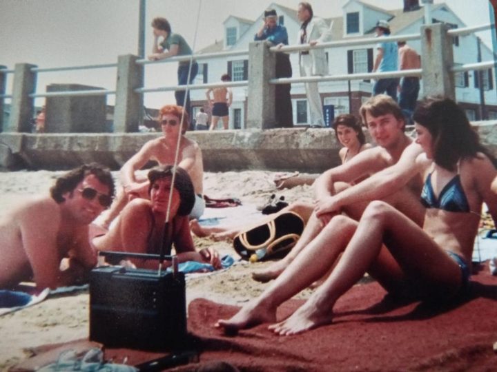 Brothers and friends on beach towels with a radio, a seawall and houses behind them