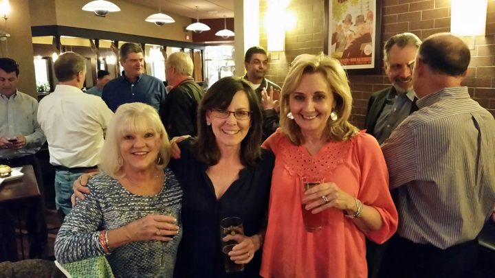 Three women smiling with drinks in a crowded brewery, brothers talking behind them