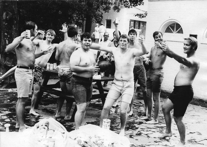 Black and white photograph of shirtless brothers raising cans on a stone patio beside a white brick building, summer 1971